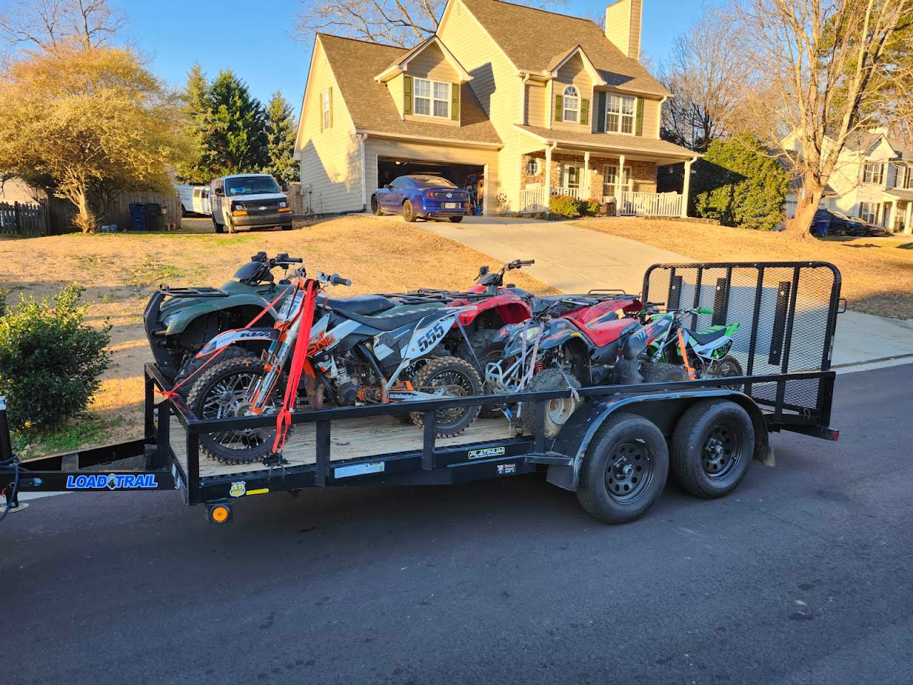 Dirt bikes and ATVs loaded on a Load Trail utility trailer in front of a home