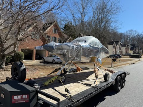 Silver aircraft fuselage on a Rent Me flatbed trailer in a neighborhood