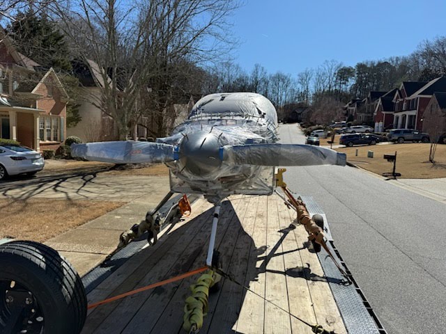Small silver aircraft fuselage with propeller loaded on a flatbed trailer