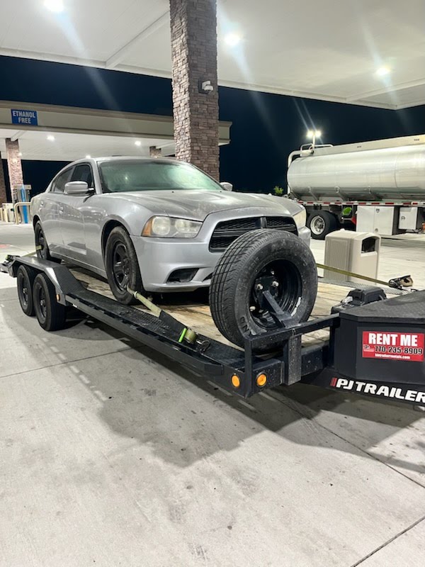 Flatbed trailer carrying a silver Dodge Charger at a gas station